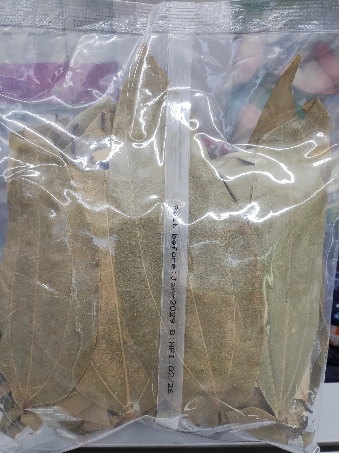 A clear plastic bag containing dried bay leaves, with a white vertical strip showing printed dates and text in the center.
