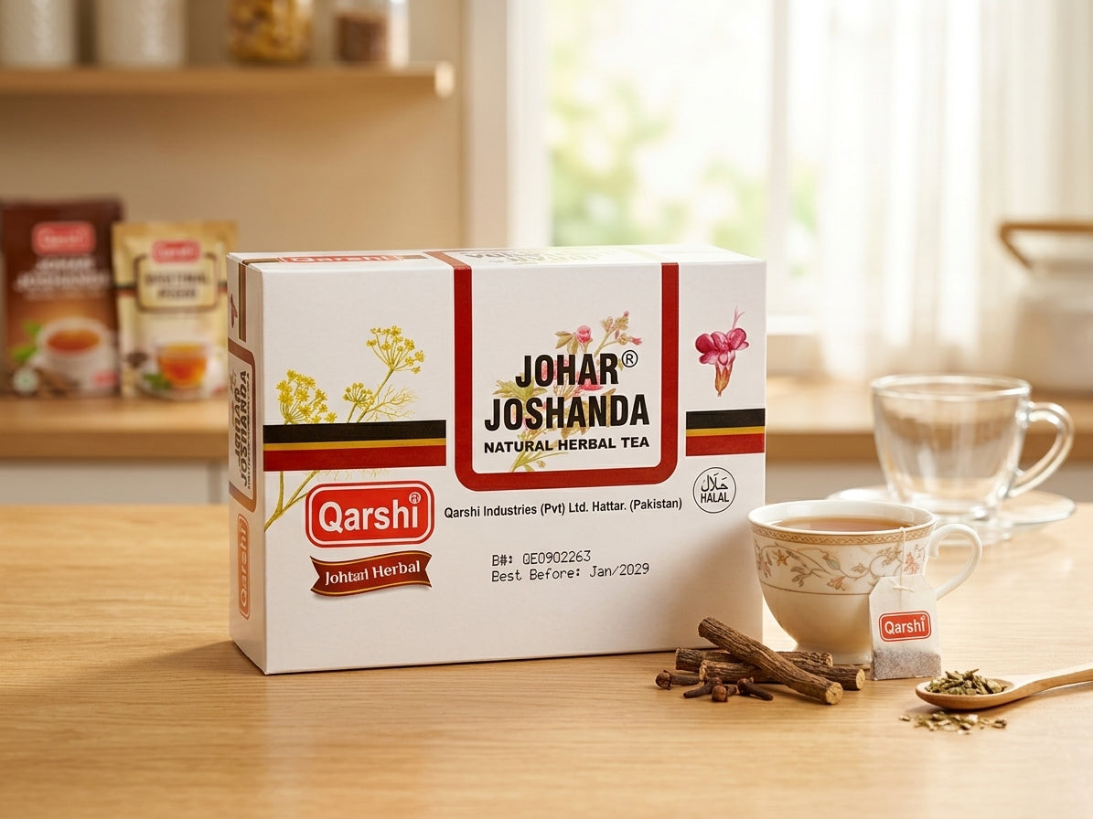 A box of Qarshi Johar Joshanda natural herbal tea sits on a wooden table, with a teacup, loose herbs, and cinnamon sticks in front of it. The background shows a softly blurred warm interior with shelves and a window.
