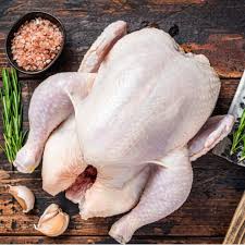 A whole raw chicken laid on a rustic wooden surface, with a small bowl of pink seasoning, rosemary sprigs, garlic cloves, and a pepper visible around it.