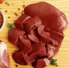 Raw red meat pieces, including a large cut piece and several smaller slices, arranged on a light wooden board with scattered peppercorns and a small bowl of seasoning.