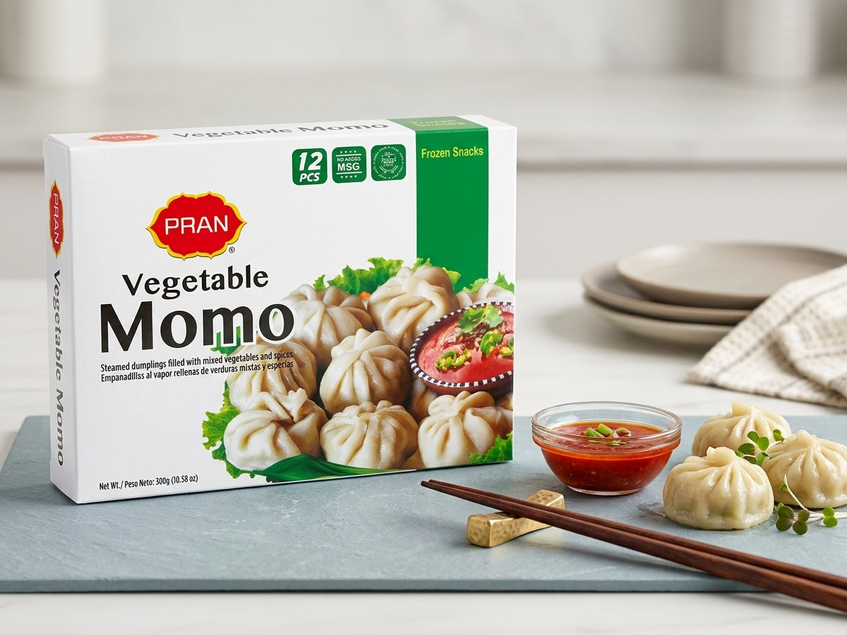 A box of PRAN Vegetable Moemo frozen dumplings sits on a gray slate board with chopsticks, a small bowl of red dipping sauce, and several dumplings on a light countertop.