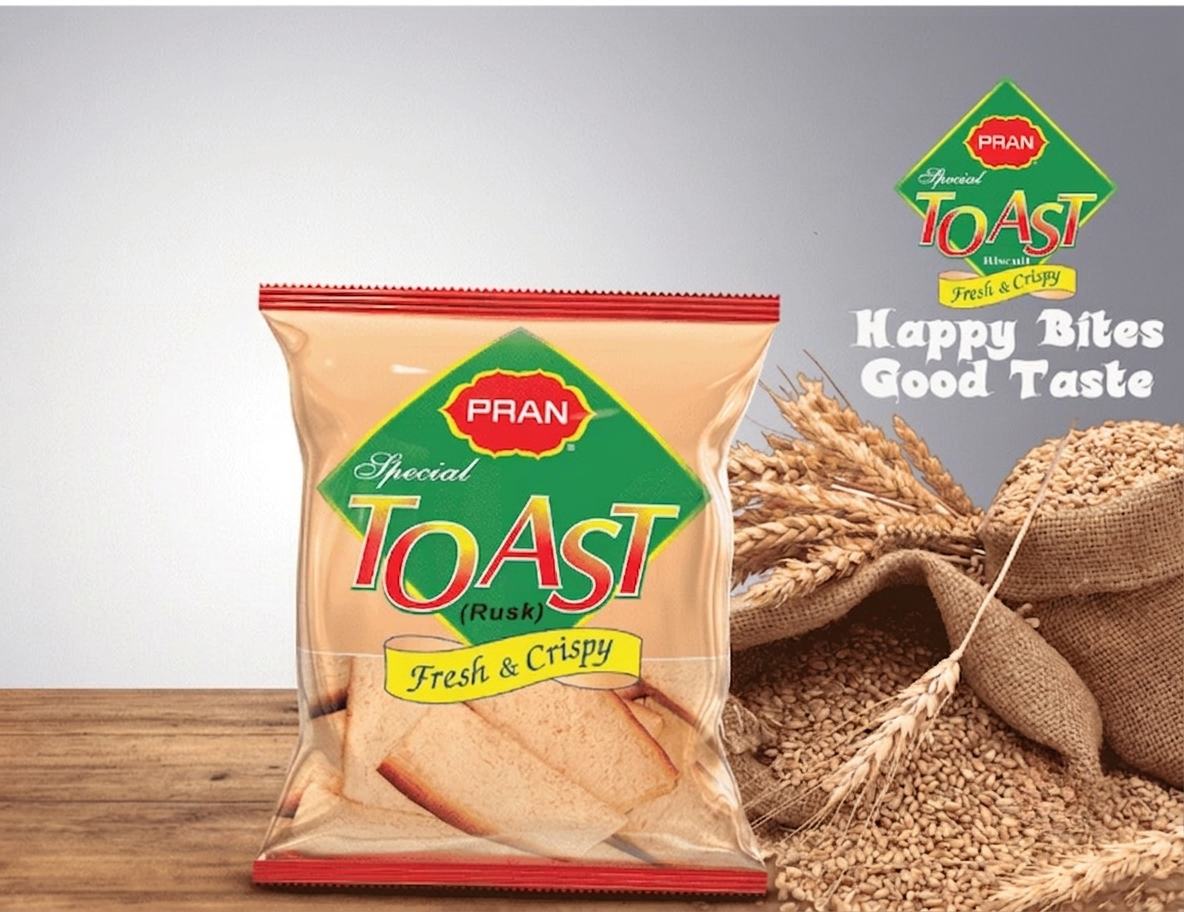 A package of PRAN Special Toast (Rusk) Fresh & Crispy, shown standing on a wooden surface next to wheat stalks and a sack of wheat grains. The package is beige with a green and red logo, and the background is a clean gray studio backdrop.