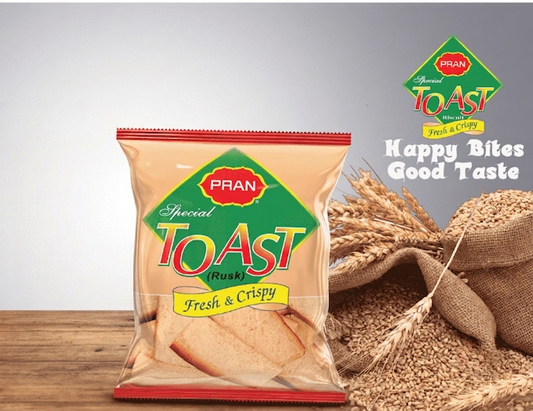 A package of PRAN Special Toast (Rusk) Fresh & Crispy, shown standing on a wooden surface next to wheat stalks and a sack of wheat grains. The package is beige with a green and red logo, and the background is a clean gray studio backdrop.