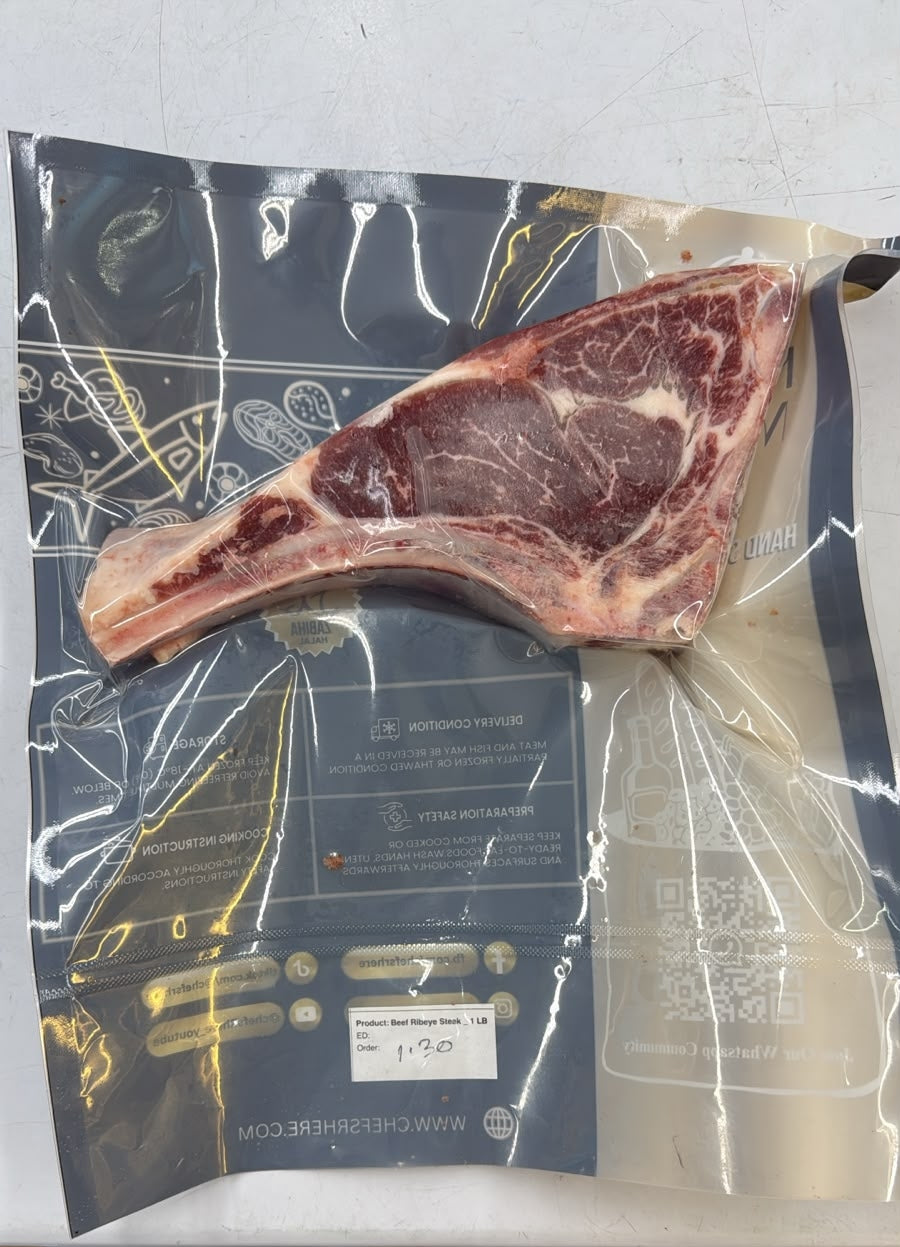 A raw bone-in steak, likely a ribeye, vacuum-sealed in a clear plastic bag with a dark blue printed label. The label features gold and white text, a QR code, and a small white price sticker.