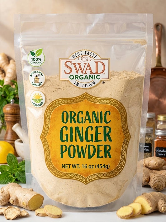 A 16 oz bag of Swad Organic Ginger Powder, shown in a clear stand-up pouch with a large yellow label. The background is styled with fresh ginger roots, mint leaves, a lemon, and small spice jars on a light surface.