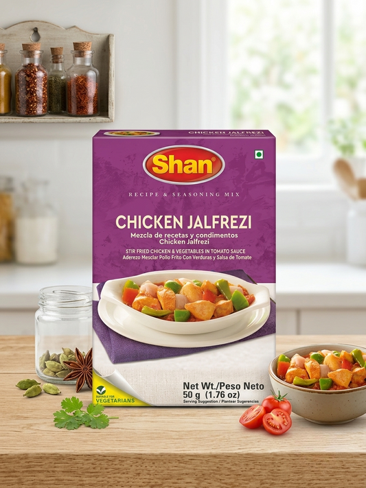 A box of Shan Chicken Jalfrezi recipe and seasoning mix displayed on a wooden countertop with a blurred kitchen background, alongside a glass jar, star anise, cilantro, and a bowl of prepared food.