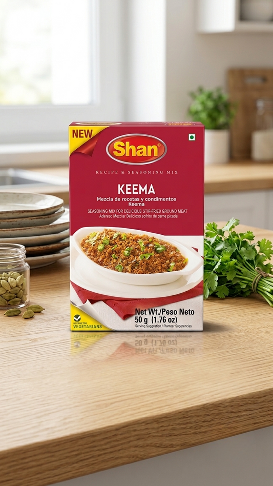 A box of Shan Keema Recipe & Seasoning Mix placed on a wooden countertop in a kitchen setting, with a bowl of food, green herbs, and kitchen items blurred in the background.