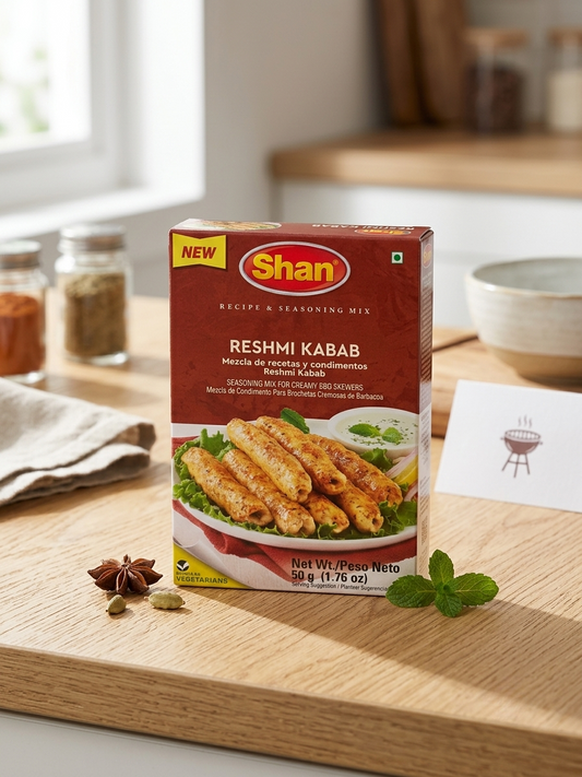 A box of Shan Reshmi Kabab seasoning mix stands on a wooden cutting board in a kitchen setting, surrounded by spices, mint leaves, and a small card.