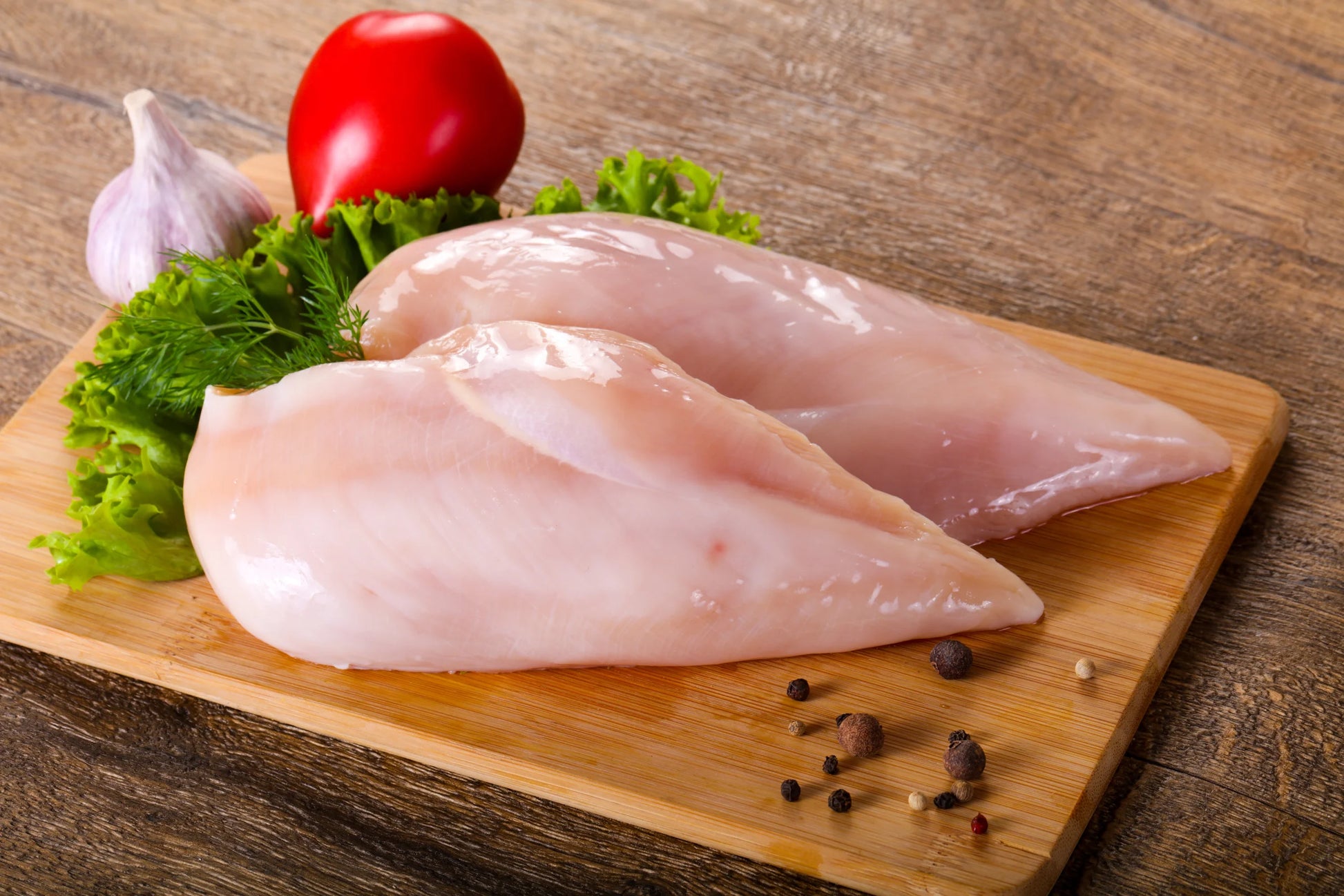Two raw  Zabiha Halal chicken breast fillets on a wooden cutting board with lettuce, a red tomato, a garlic bulb, and scattered peppercorns on a rustic wooden table.