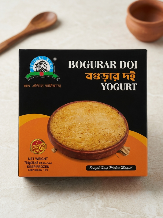 A box of Boogar Doi Yogurt on a light textured surface, with a wooden spoon and clay bowl blurred in the background. The packaging is black and orange with a photo of a baked yogurt dish.