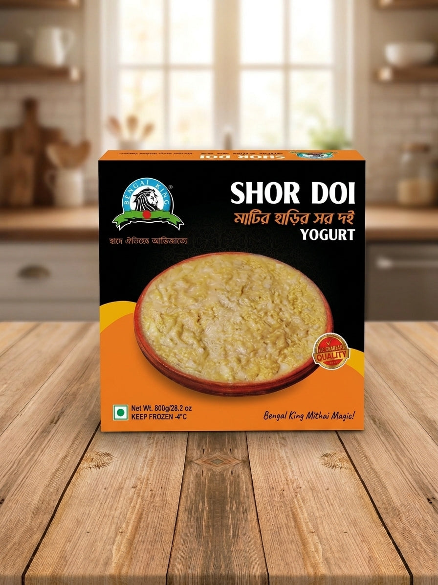 A box of Shor Doi Yogurt (frozen yogurt) by the brand Shor, placed on a wooden table with a blurred kitchen background. The packaging is black and orange with a photo of the yellowish-cream yogurt on the front.