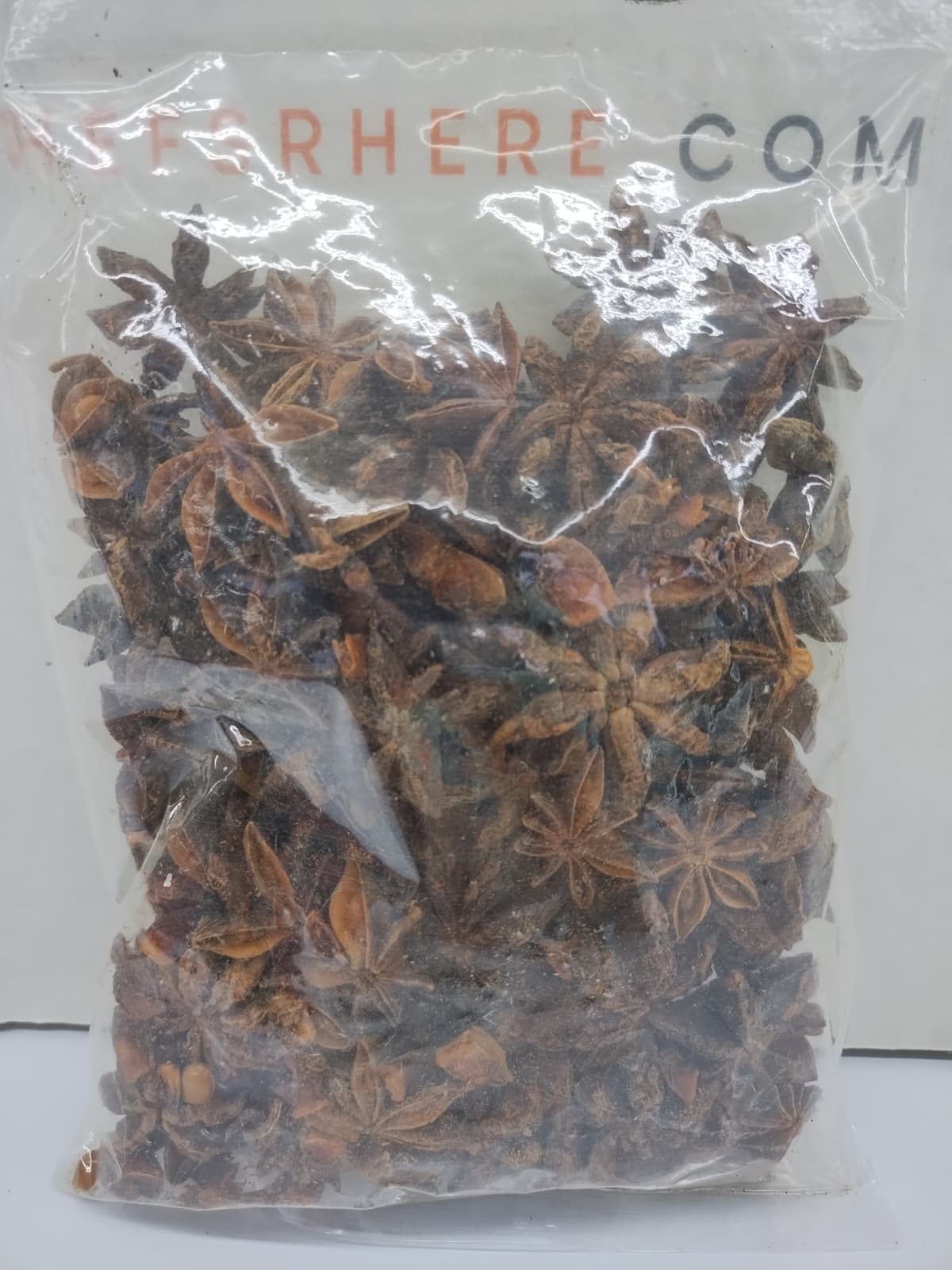 A clear plastic bag filled with dried star anise spices, with a visible 'STARHERE.COM' watermark at the top, set against a plain white background.