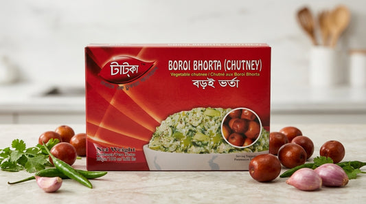 A box of Tatka Boroi Bhorta (Chutney) placed on a light countertop with whole red chilies, garlic, and cilantro arranged around it. The background shows a softly blurred kitchen setting with wooden spoons in a container.