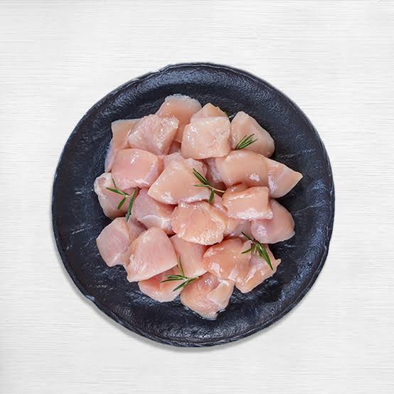 Raw zabiha halal chicken cube pieces from boneless chicken breast on a dark plate with rosemary on a white background